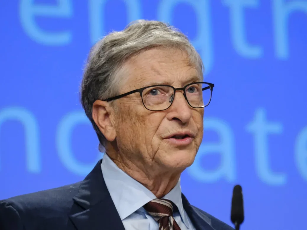 Bill Gates wearing a suit and tie, speaking into a microphone with a blue stage background behind him