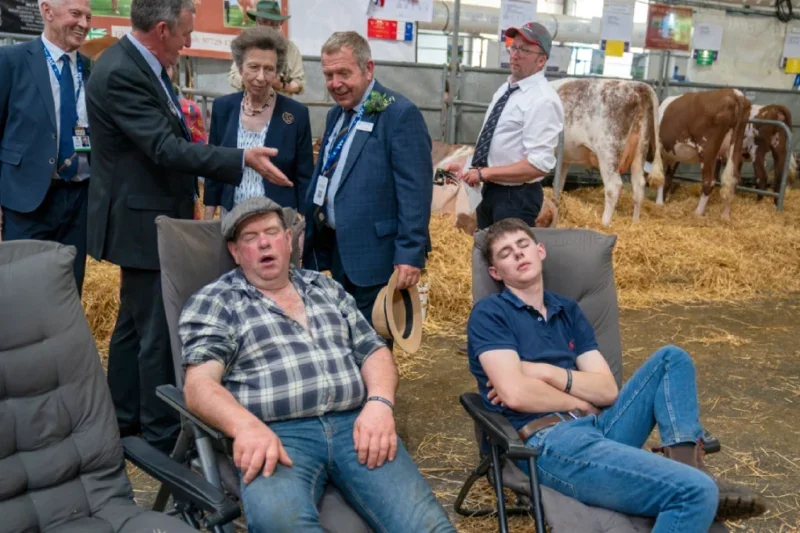 Two farmers sleeping on chairs in cattle stalls with The Princess Royal walking behind them