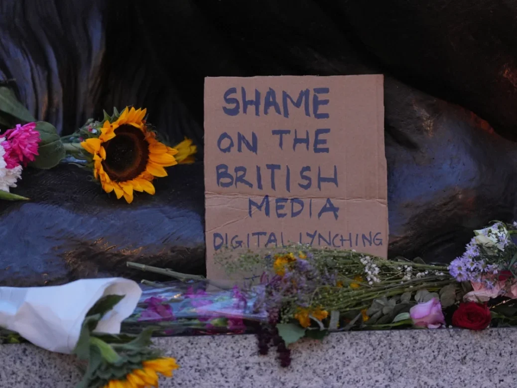 Flowers on the floor with sign written on cardboard saying 'Shame on the British media. Digital lynching'