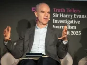 New York Times executive editor Joe Kahn appears on stage at the Sir Harry Evans Investigative Journalism Summit in London in 2025. Kahn, wearing a dark grey blazer over a blue shirt undone at the top button, is seen mid-speech, gesturing with his arms apart.