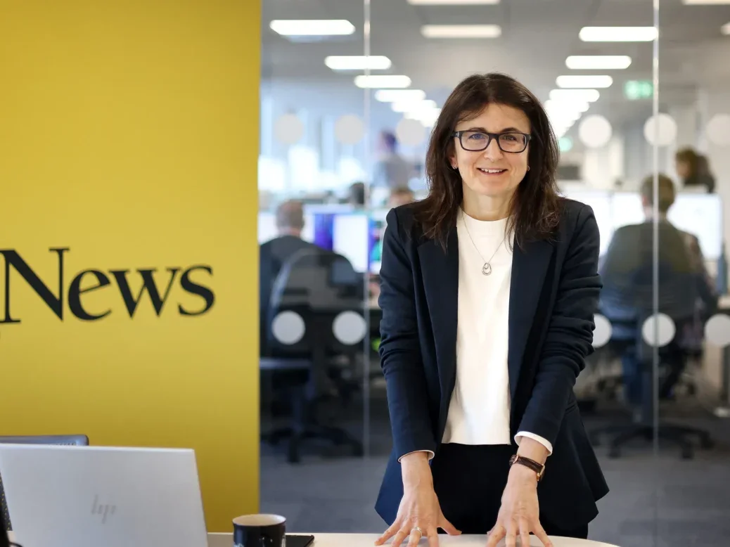 Manchester Evening News editor Sarah Lester pictured in a meeting room with the newsroom behind her, smiling at the camera