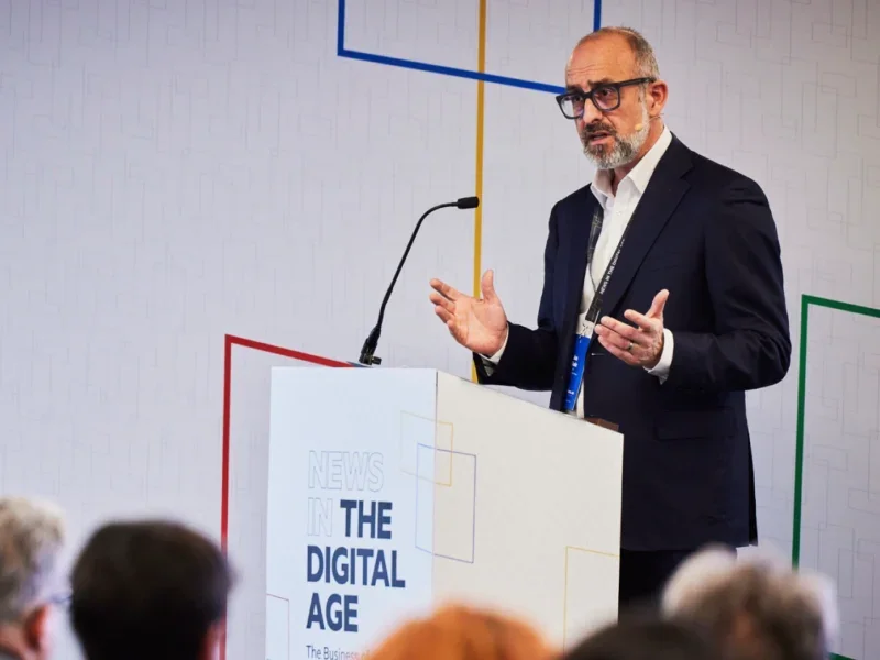 Jon Slade, Financial Times CEO, speaking at a lectern with News in the Digital Age branding with audience heads in foreground