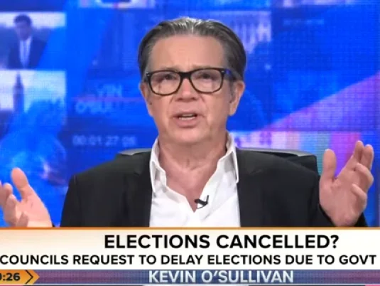 Kevin O'Sullivan gesturing with both hands while talking at camera with blue studio background behind him. Ticker on screen shows Talk logo, time of 19.26 and strapline: 'Elections cancelled? Councils request to delay elections due to govt shake up'