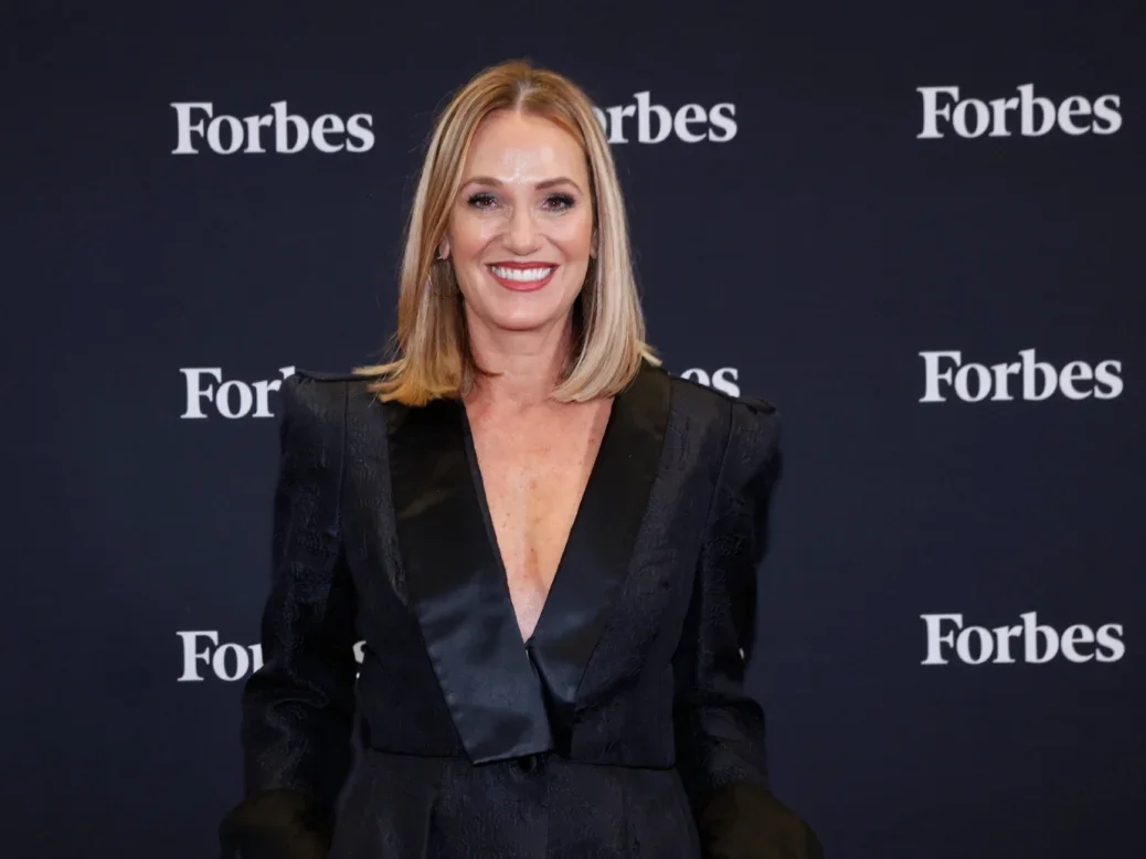 Sherry Phillips, Forbes CEO stands wearing black blazer in front of black backdrop with Forbes logo on it several times